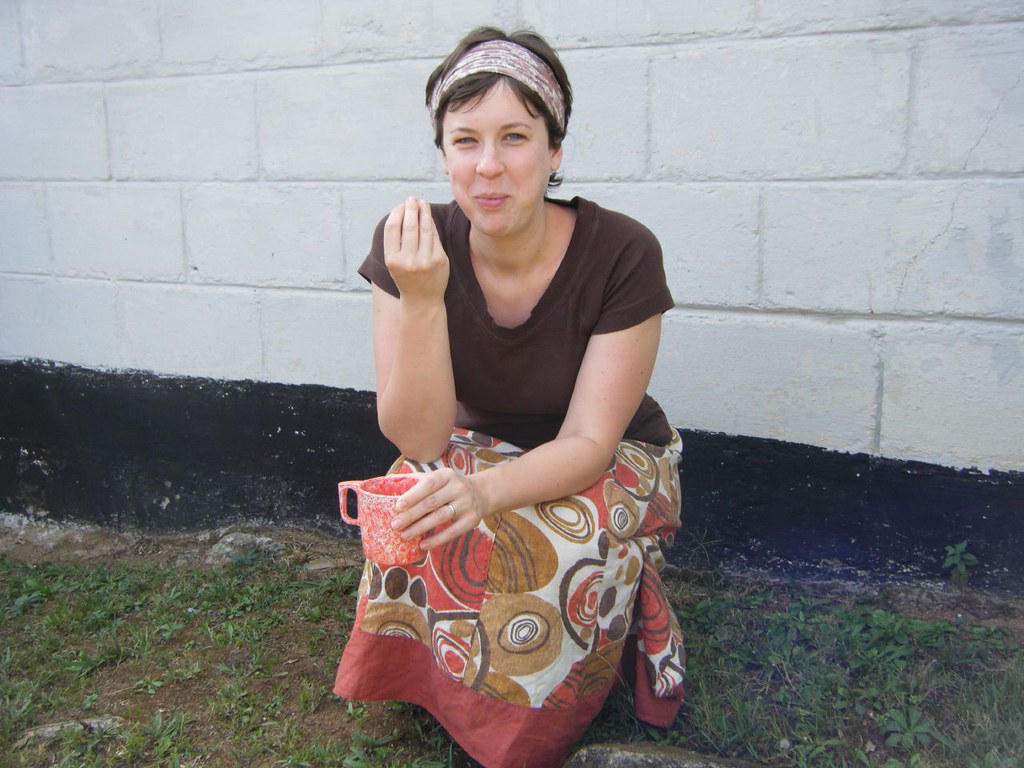 A much younger Virginia Spielmann crouching down with a cup of tea in her hand. She is wearing a head scarf and skirt and tshirt and eating mandazi and drinking chai.