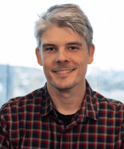 A man with short gray hair smiling at the camera, wearing a red and navy plaid button-down shirt, photographed against a blurred window background.