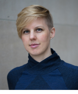 A woman with short blonde hair, undercut on one side, wearing a dark navy turtleneck sweater, photographed against a gray textured background.