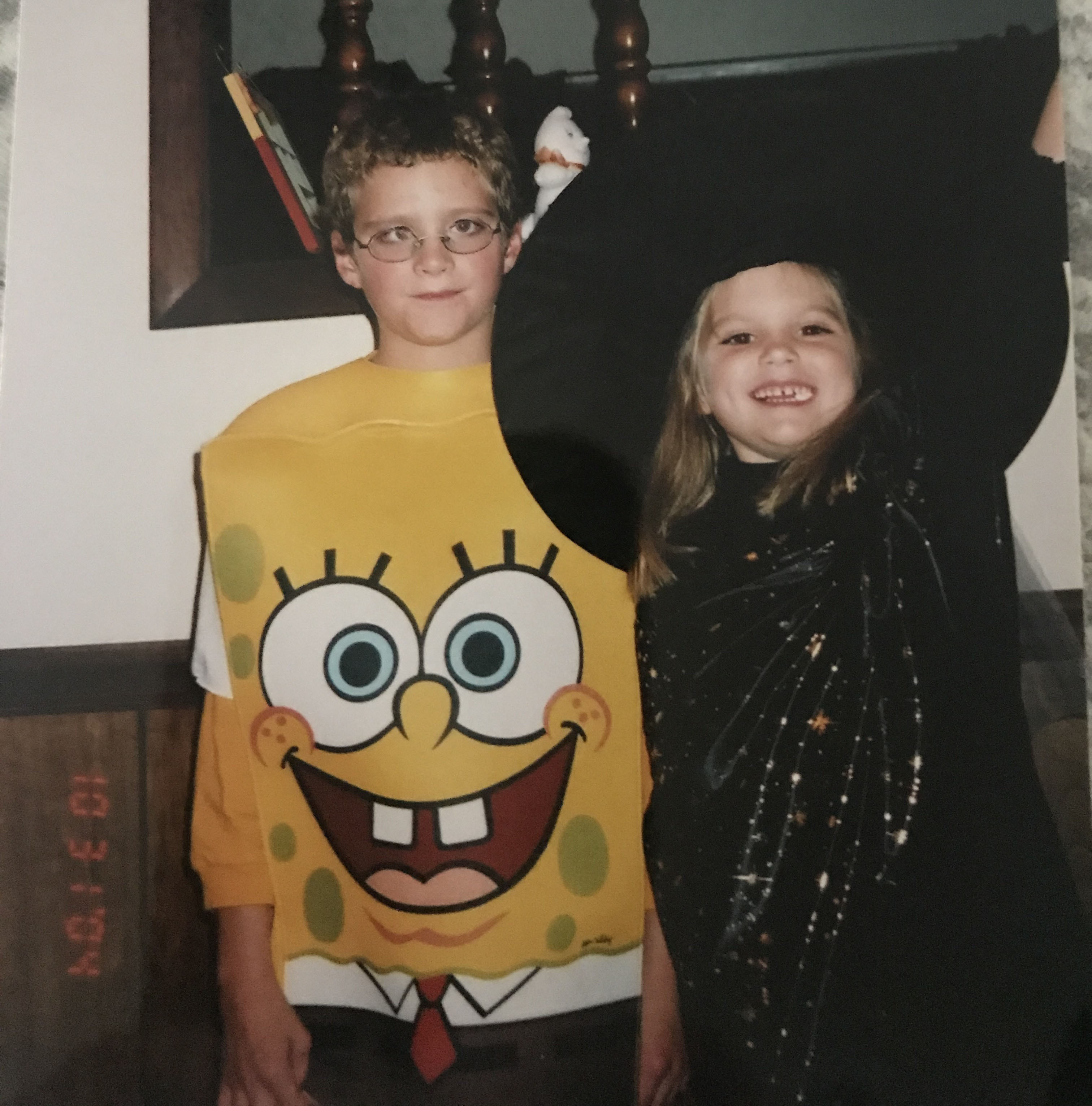 A childhood photo of a boy and girl in Halloween costumes. The boy is wearing glasses and a SpongeBob SquarePants costume, while the girl is dressed as a witch in a black outfit with silver star patterns, smiling with one arm raised. They are standing indoors in front of a wooden banister and a mirror.