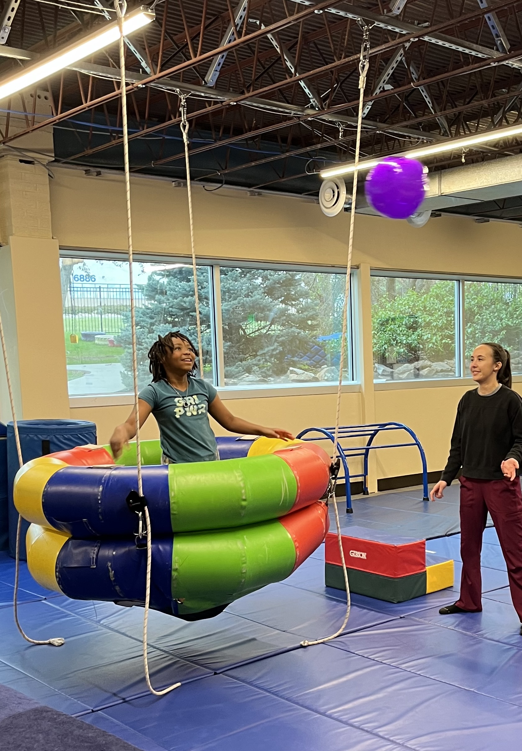 Photo of a young African American girl standing in a swing looking up at a ball headed towards her. A Hispanic female occupational therapist stands alongside her.