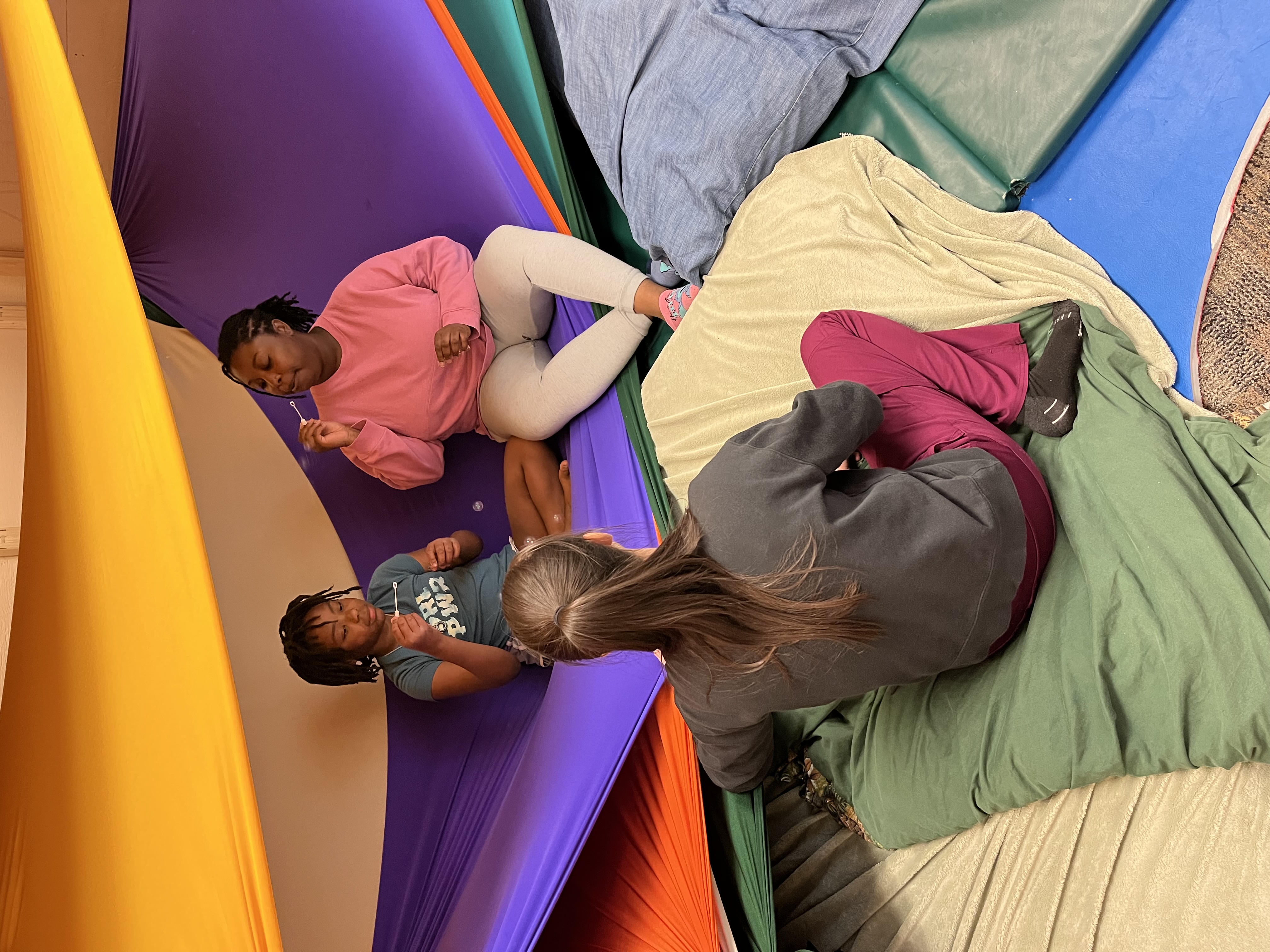 A young African American girl sits on Lycra next to an adult African American female on Lycra they are blowing bubbles together. An adult sits outside the Lycra with their back to the camera their hair is in a ponytail.