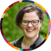 A smiling person with short dark hair featuring purple highlights, wearing glasses and a dark blazer. They are photographed outdoors in front of a lush green background, framed by a colorful circular border.