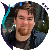 Circular headshot of a man with short brown hair and a red beard, smiling at the camera. He's wearing a dark button-up shirt over a gray t-shirt, photographed outdoors at night with blurred city lights in the background. A teal and purple abstract logo mark appears to the left of the frame.