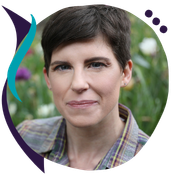 Headshot of Coriander Woodland, a person with short dark hair, wearing a plaid shirt, with flowers in the background.