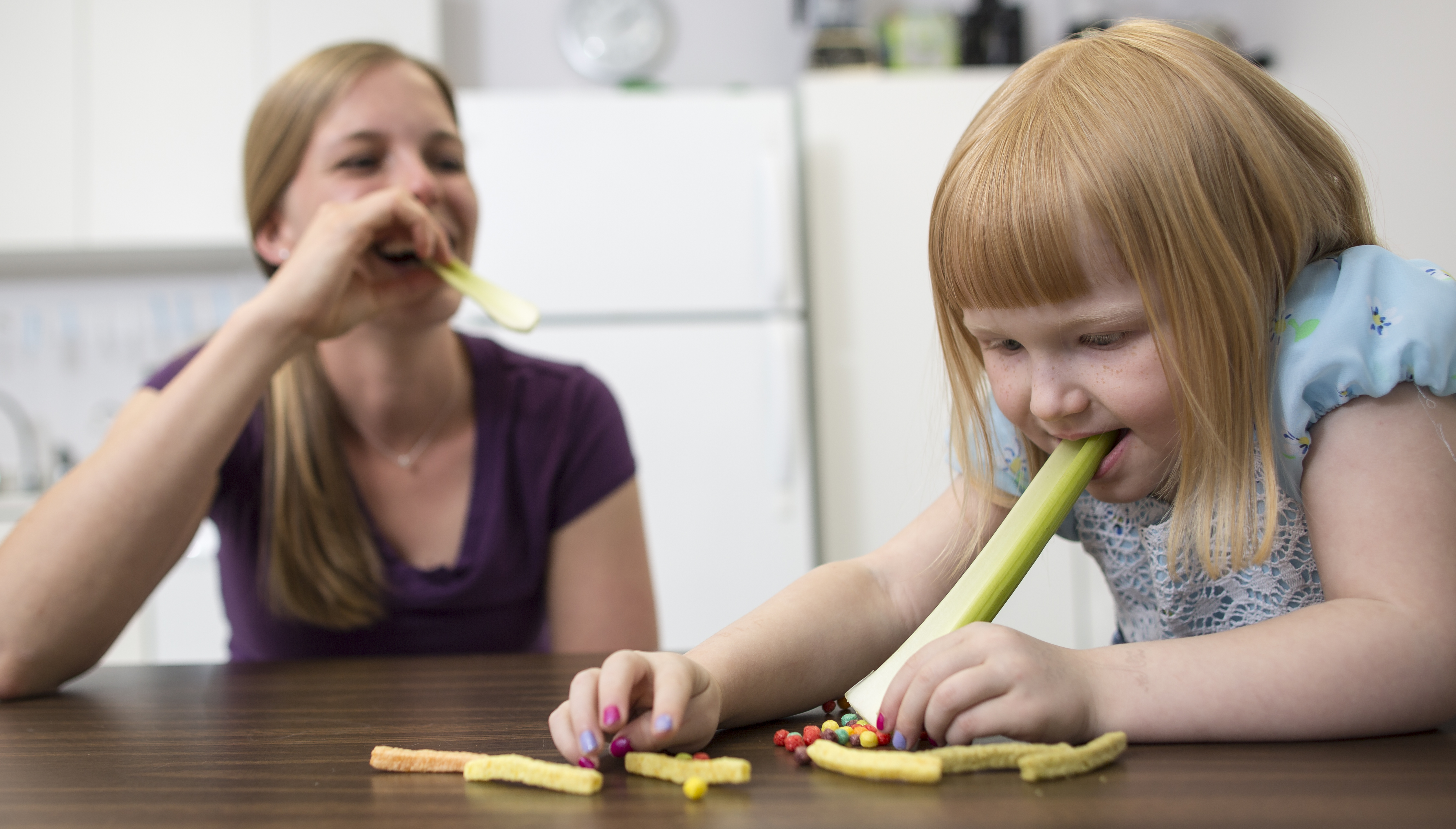 Child is sitting at the table with a long piece of celery in her mouth with the end touching the table. There are candy pieces and veggie straws on the table around her. A therapist is sitting next to her with a piece of celery in her mouth