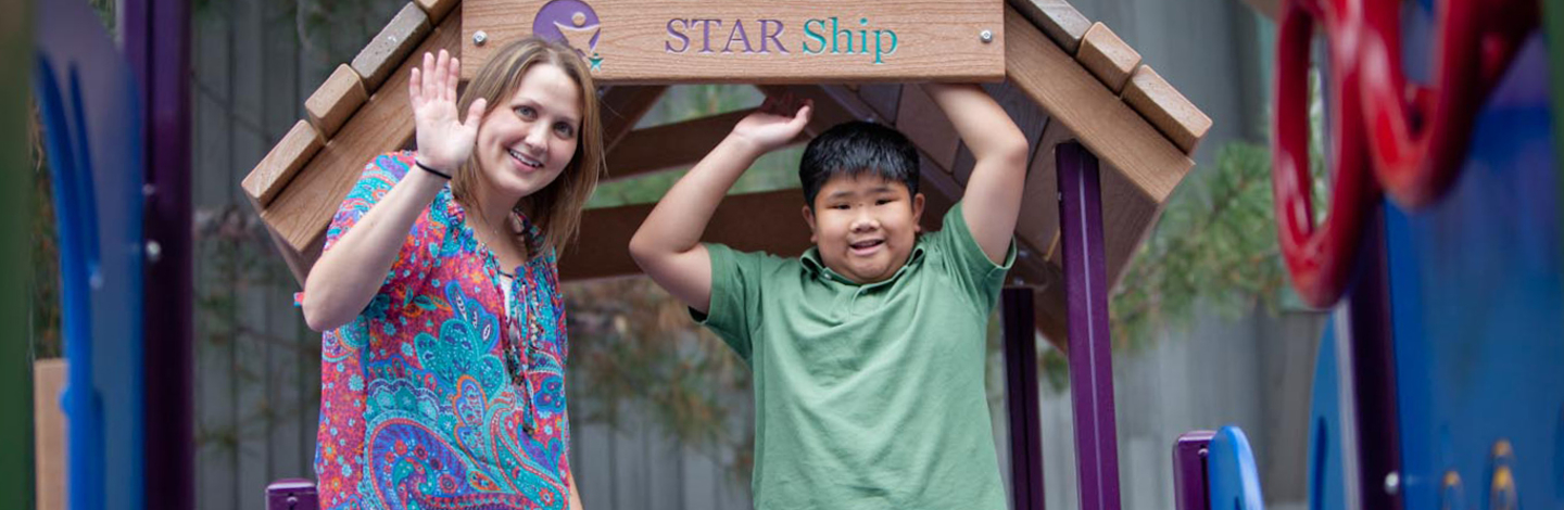 Child standing under a sign that says "Star Ship" smiling at the camera next to a therapist waving at the camera