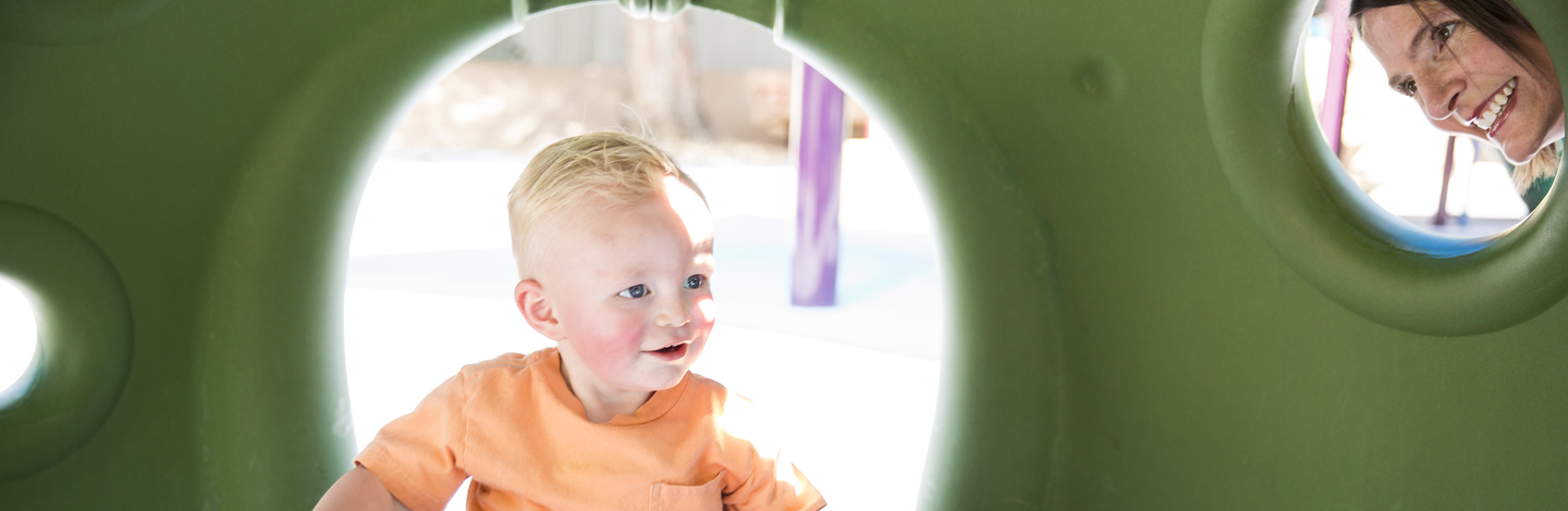 Young child playing on playground equipment while an adult peers at him smiling.