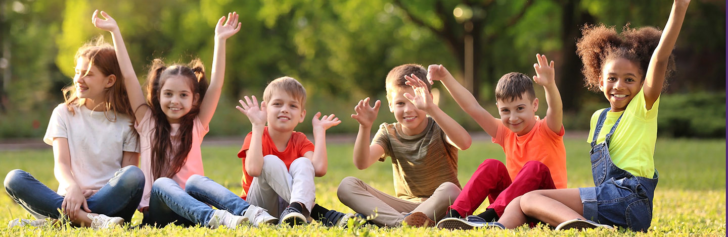 Six children sitting in the grass next to one another with their hands in the air, smiling at the camera