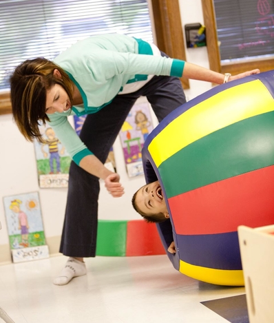 A child is inside a colorful play barrel with his head sticking out smiling as an adult holds the barrel looking at the child with excitement