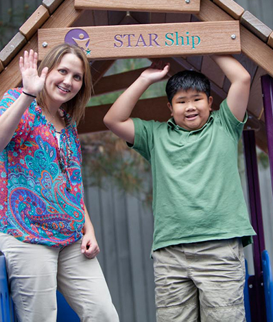 Child standing under a sign that says "Star Ship" smiling at the camera next to a therapist waving at the camera