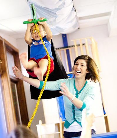 Child is using a zipline while the therapist has their arms out to support them in case they fall