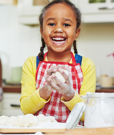 Young child is standing at the kitchen counter with flour and dough in front of her. She is smiling while holding ball of dough in her hands and flour on her face