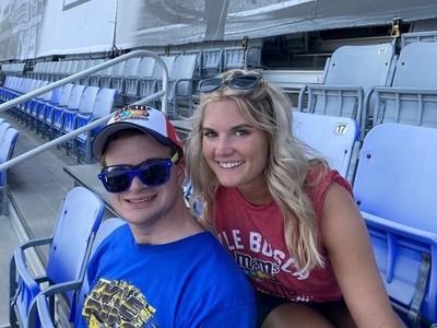 A man and woman smiling while seated in a stadium. The man is wearing a colorful cap, blue sunglasses, and a bright blue Kyle Busch racing T-shirt. The woman, with long blonde hair, is wearing a red sleeveless top and has sunglasses on her head. Empty blue and gray stadium seats surround them.