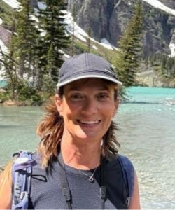 A person smiling outdoors, wearing a gray baseball cap and sleeveless athletic top with a backpack strap visible. A turquoise alpine lake and evergreen trees are visible in the background.