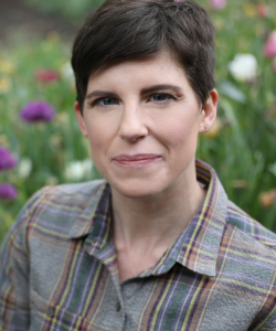 Headshot of Coriander Woodland, a woman with short dark hair wearing a plaid shirt, photographed outdoors in front of colorful garden flowers.
