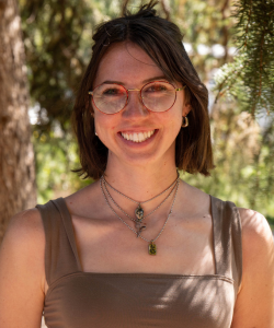 Portrait of a smiling fair skinned woman with short brown hair and glasses, wearing a brown top and layered necklaces, standing outdoors in front of greenery.
