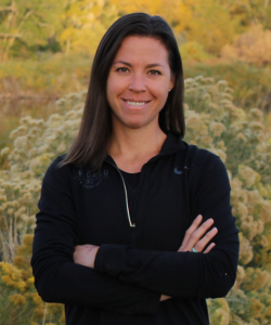 A woman with long dark brown hair stands outdoors in front of tall grasses and yellow wildflowers. She is wearing a black zip-up jacket and smiling with her arms crossed. The background shows a natural, sunlit landscape.