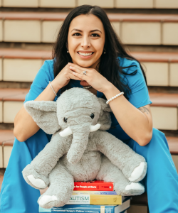 A smiling woman in blue scrubs sits on outdoor steps, resting her chin on her hands. She has long dark hair and is seated behind a plush gray elephant, which is placed on a stack of books about autism and child development.